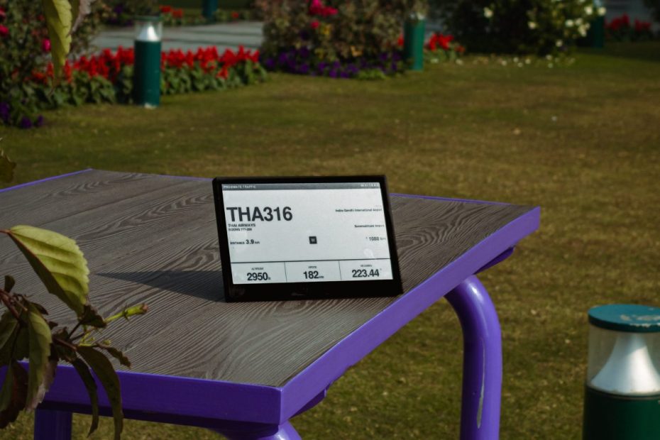TRMNL e-ink display showing a flight number and stats, standing on a purple outdoor table in a garden
