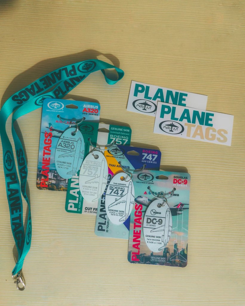 Four PlaneTags aircraft-aluminum tags on their cards (Air Canada A320, Boeing 757, Thai Airways 747, Delta DC-9) with a teal PlaneTags lanyard and two stickers on a wooden table
