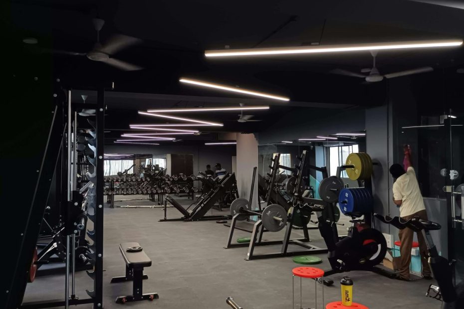 Dark gym floor with weight racks, benches and a leg press under strip lights, with one person at the back